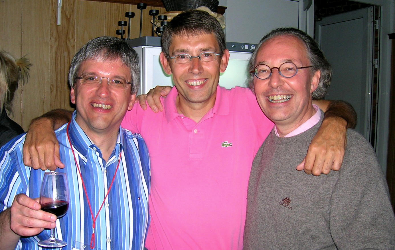 Trois hommes souriants, bras dessus dessous; l'homme de gauche tient un verre de vin rouge, au milieu porte un polo rose et à droite un pull gris.