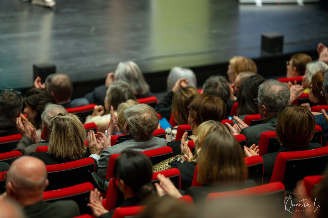 Public assis dans des sièges rouges dans un théâtre, applaudissant en direction de la scène.