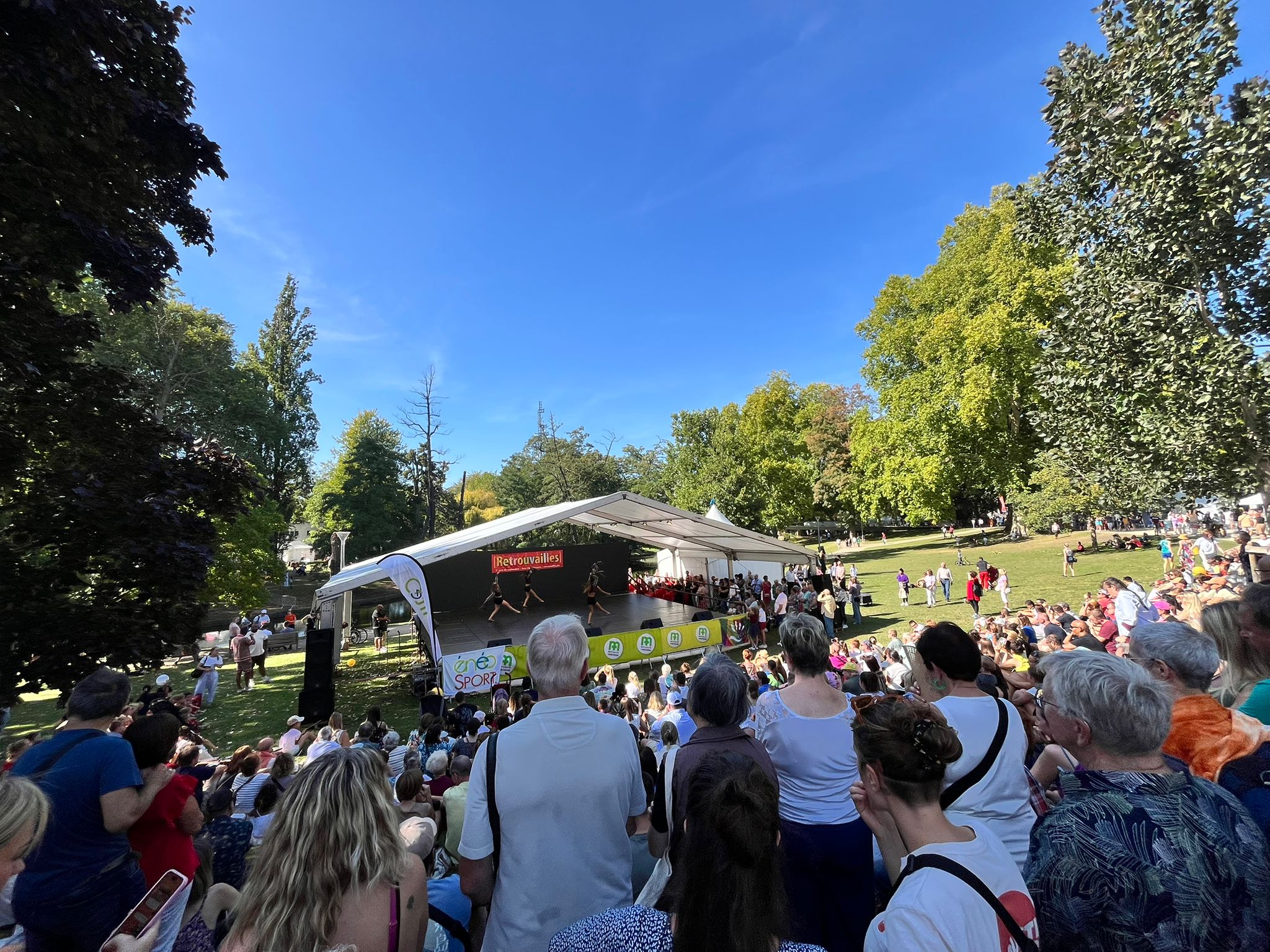 Foule assistant à une performance sur une scène couverte dans un parc ensoleillé avec des arbres verts en arrière-plan Parc de la Boverie