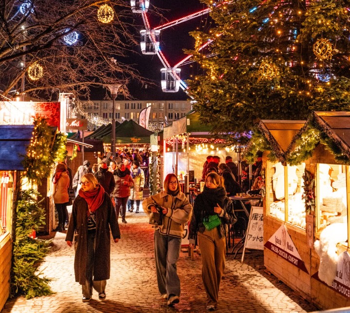 Foule se promenant dans un marché de Noël éclairé la nuit avec des cabanes décorées, un grand sapin et une grande roue en arrière-plan.