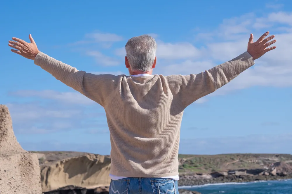 Hombre mayor de espaldas con los brazos levantados mirando el horizonte