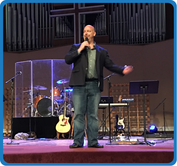 Man speaking on stage in a church with musical instruments and a large wooden cross in the background.