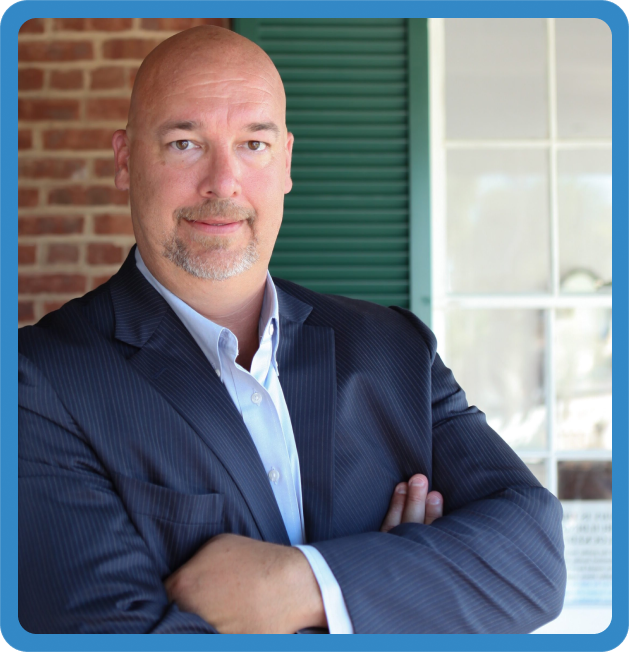 Bald man with a goatee wearing a dark blue pinstripe suit and light blue shirt standing with arms crossed in front of a green shutter and brick wall.
