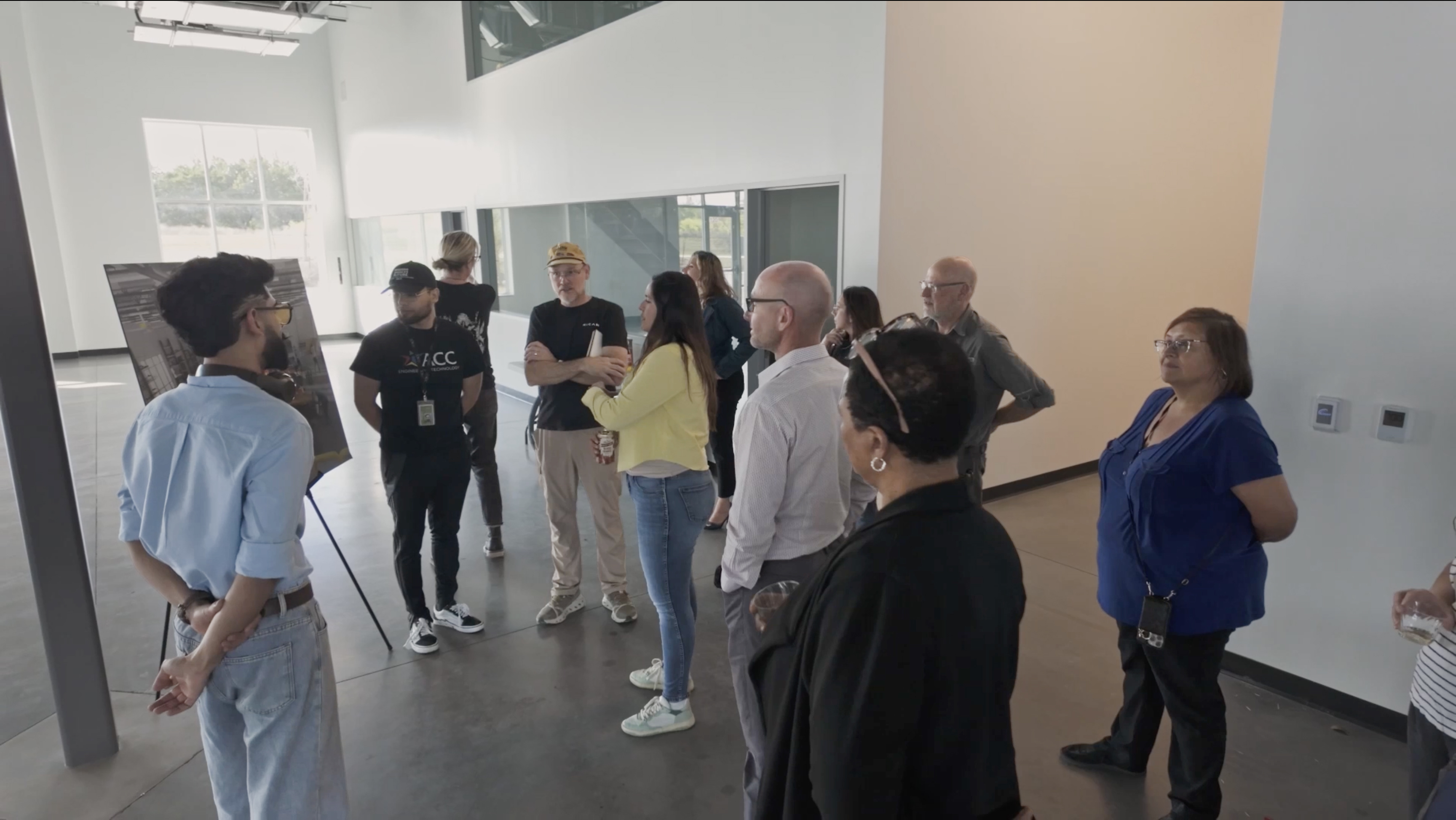 A group of people gathers in a spacious, modern room with large windows. One person is presenting in front of a display board. The atmosphere is attentive and engaged.