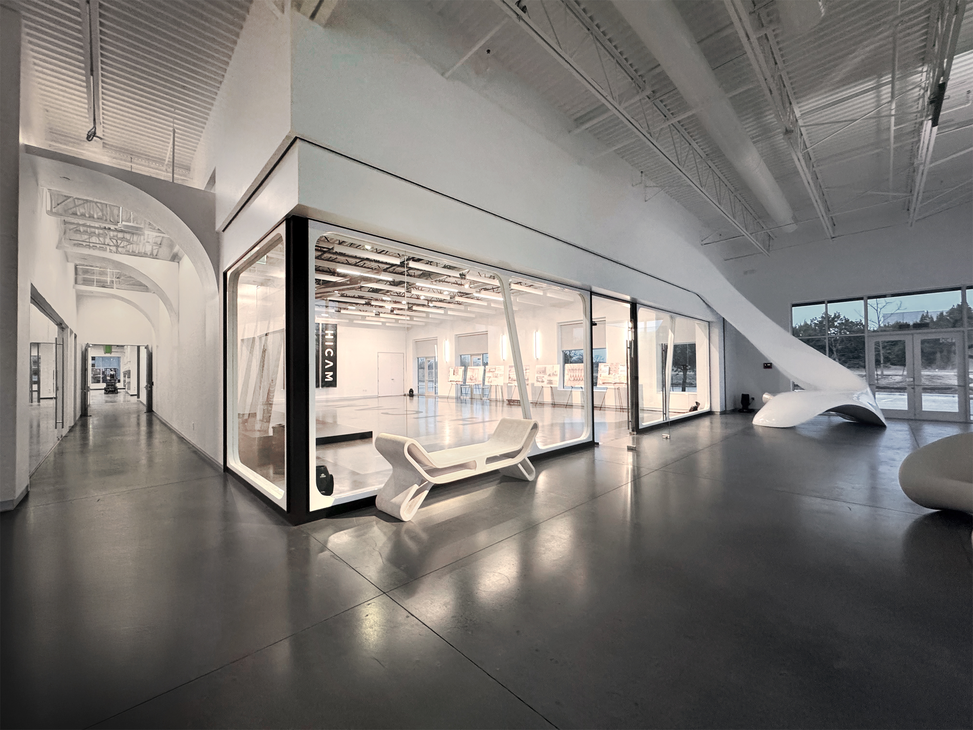 HICAM’s modern, spacious lobby with high ceilings, polished concrete floors, and sleek white architectural features. A large glass-walled event space room is centrally positioned, surrounded by sculptural furniture and natural light from large windows.