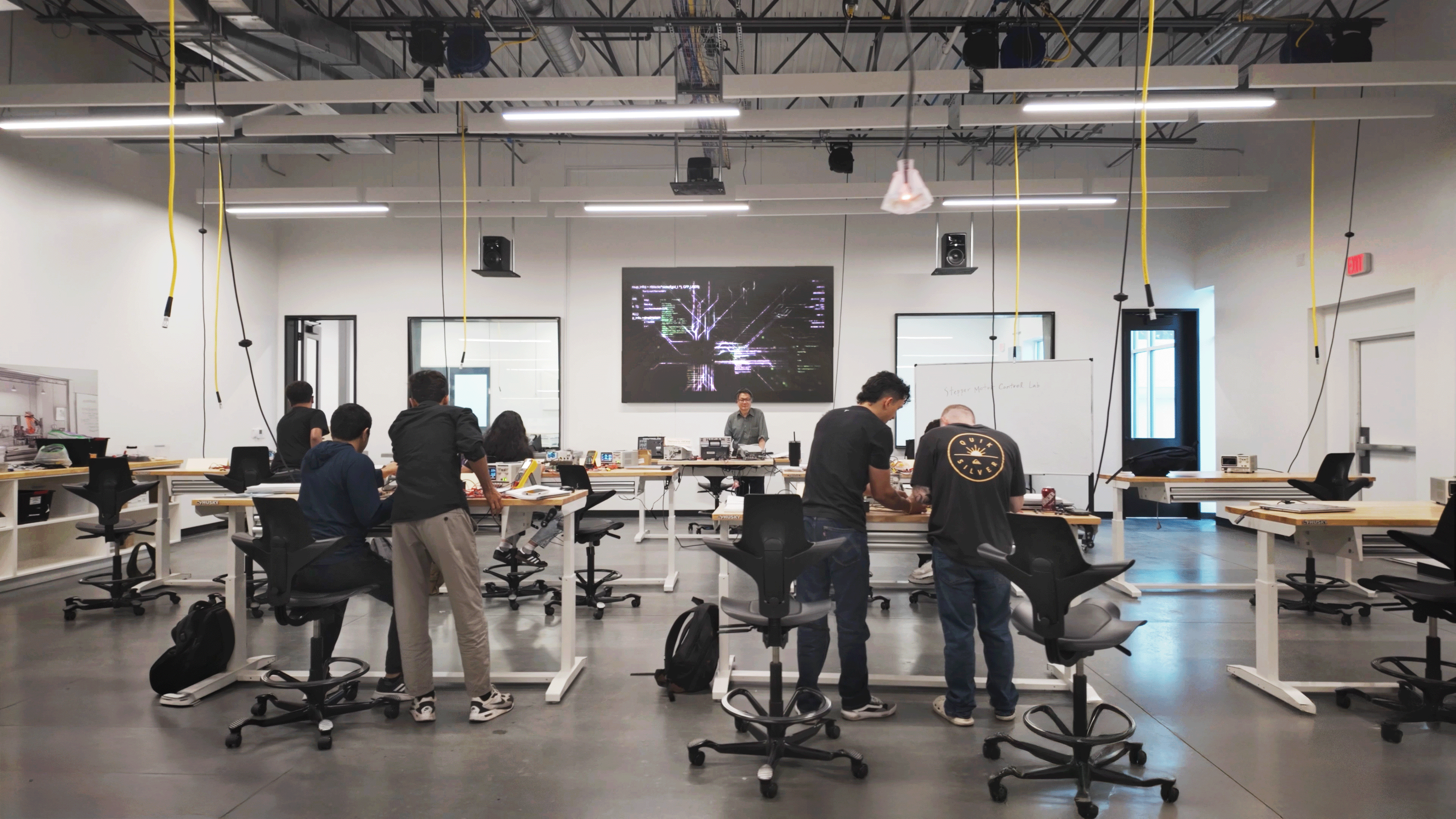 A spacious tech classroom with group of engineering students actively engaged in experiential learning. A large screen displays graphics. The atmosphere is collaborative and focused.