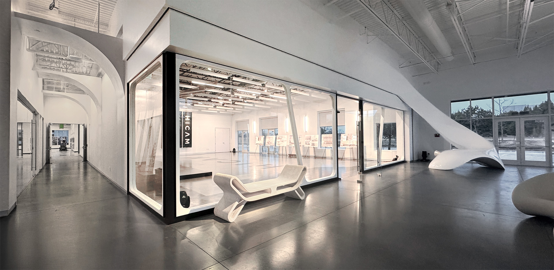 HICAM’s modern, spacious lobby with high ceilings, polished concrete floors, and sleek white architectural features. A large glass-walled event space room is centrally positioned, surrounded by sculptural furniture and natural light from large windows.