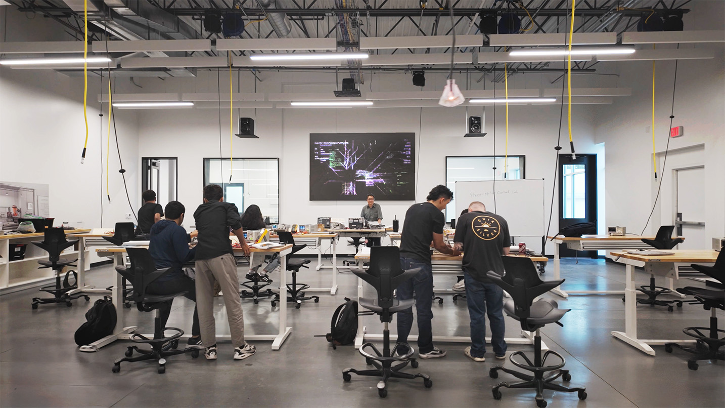 A modern classroom with students working at standing desks. A large screen shows a diagram, and the instructor observes. The atmosphere is focused and collaborative.