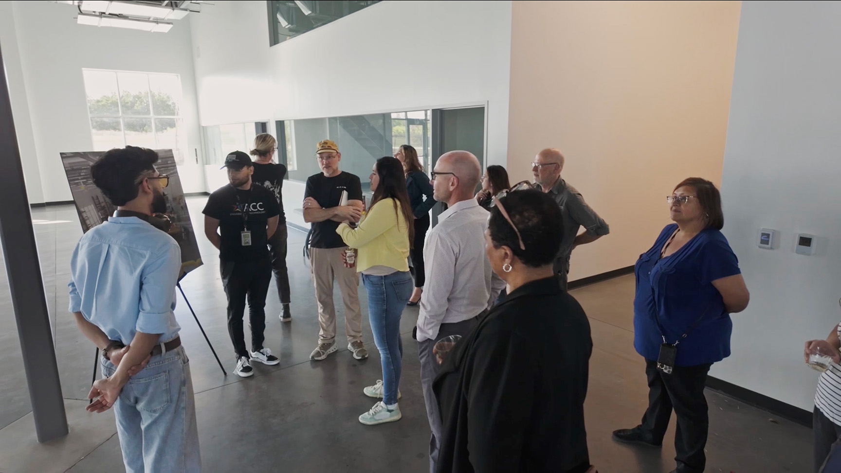 A group of people gathers in a modern, well-lit room with large windows. A presenter with a blue shirt addresses them. The atmosphere is attentive and engaged.