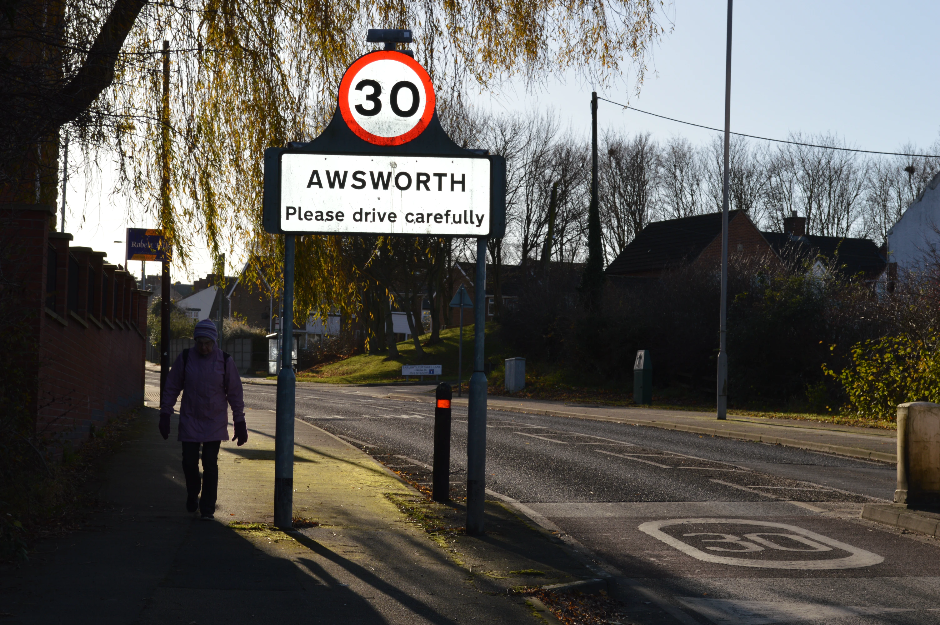 Road sign displaying a 30 speed limit above the word AWSWORTH and the message Please drive carefully, with a pedestrian walking on the sidewalk beside it.