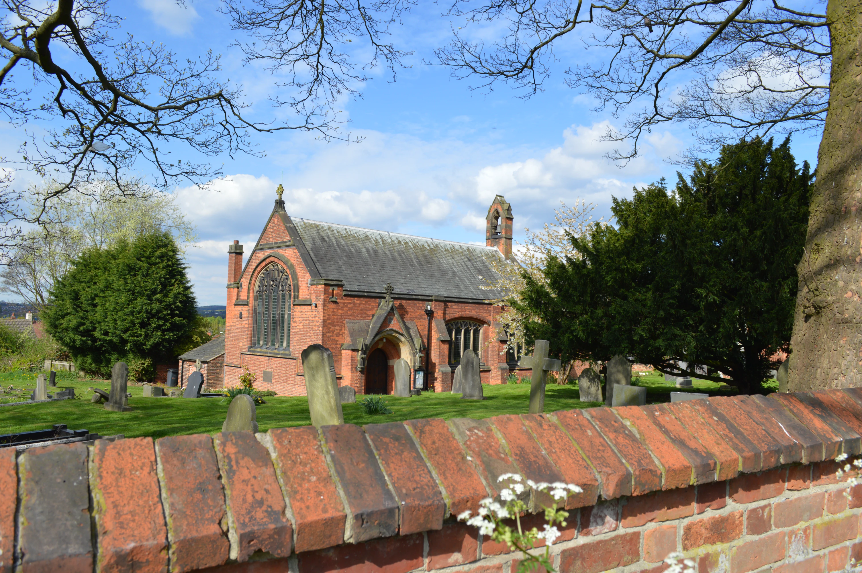Red brick church with stained glass windows seen over a brick wall, surrounded by green grass, gravestones, and trees under a blue sky.