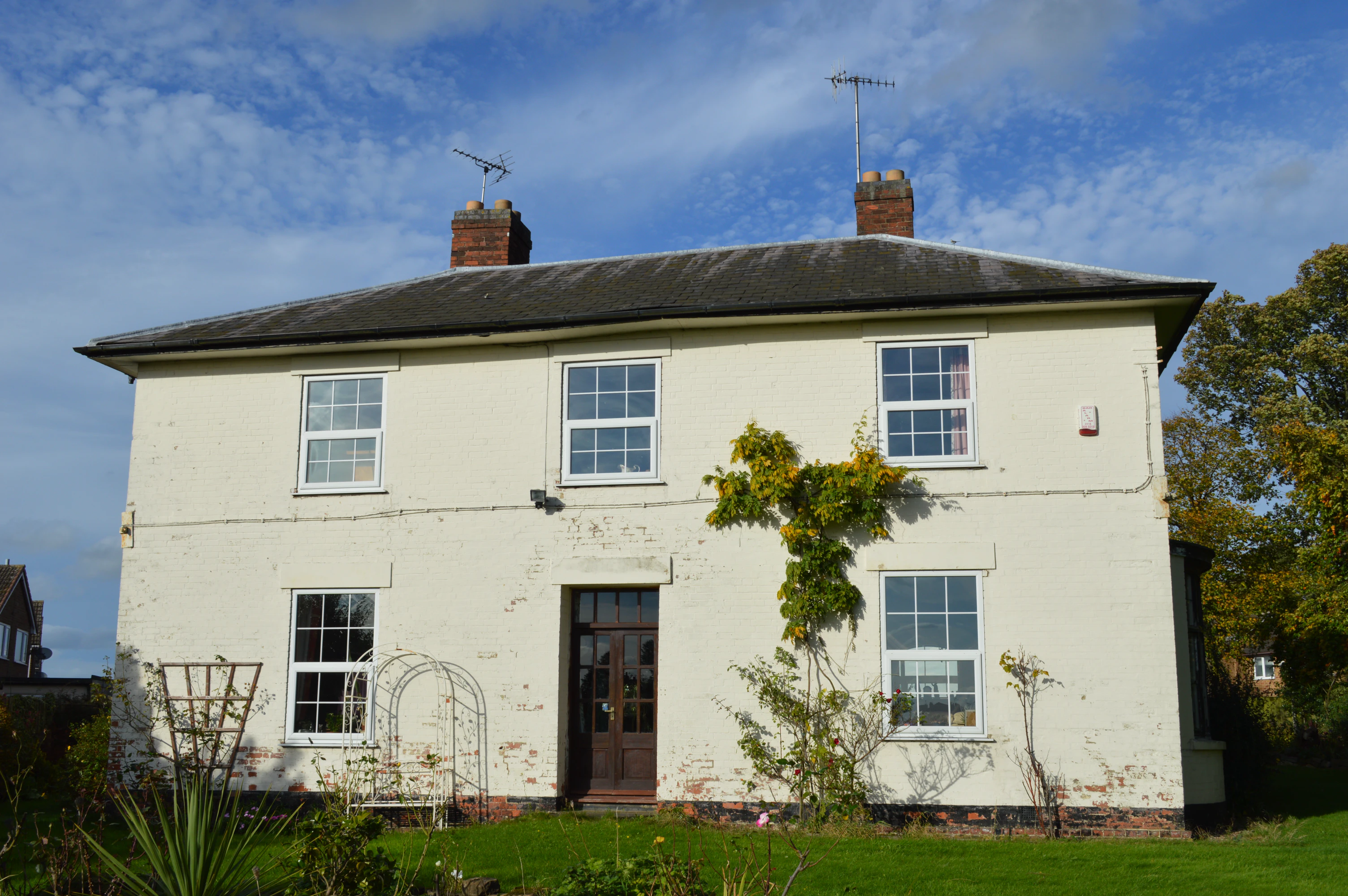 Two-story brick house painted white with a dark roof, two chimneys, and five windows, surrounded by a green lawn and garden under a partly cloudy blue sky.