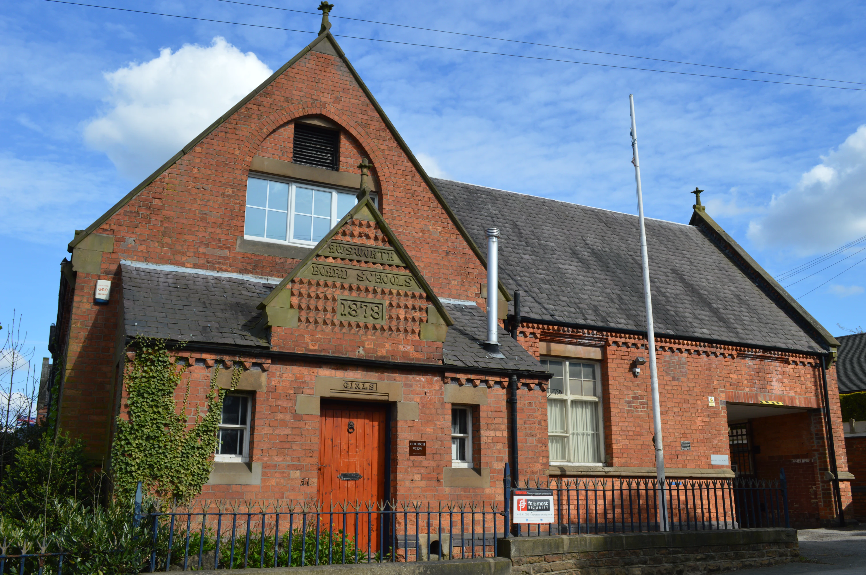 Brick building with a steep slate roof and red wooden door under the sign 'Awsworth Board Schools 1878' on a sunny day.