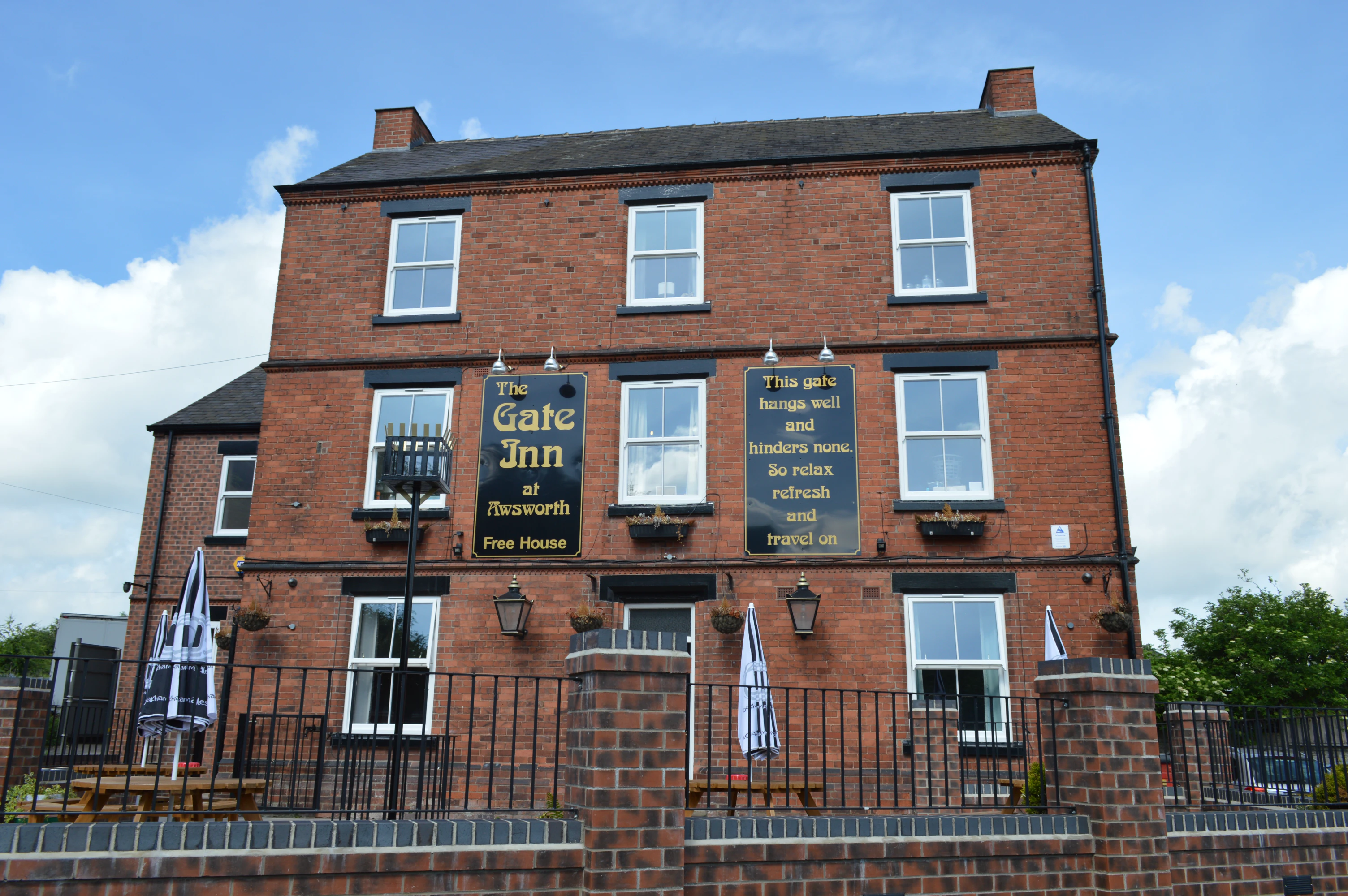 Red brick three-story building with black railings and two large signs reading 'The Gate Inn at Awsworth Free House' and a welcoming poem.