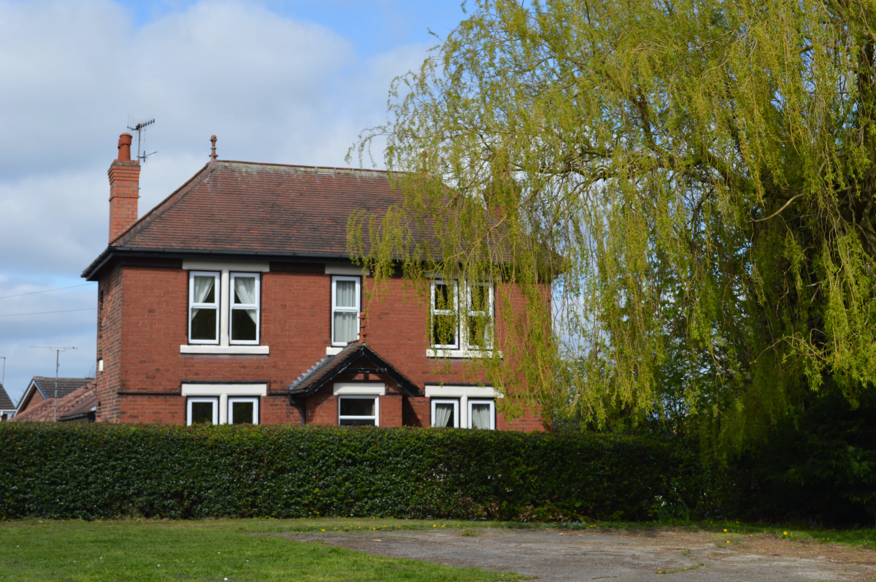 Two-story red brick house with white-framed windows behind a hedge and large tree branches on the right.