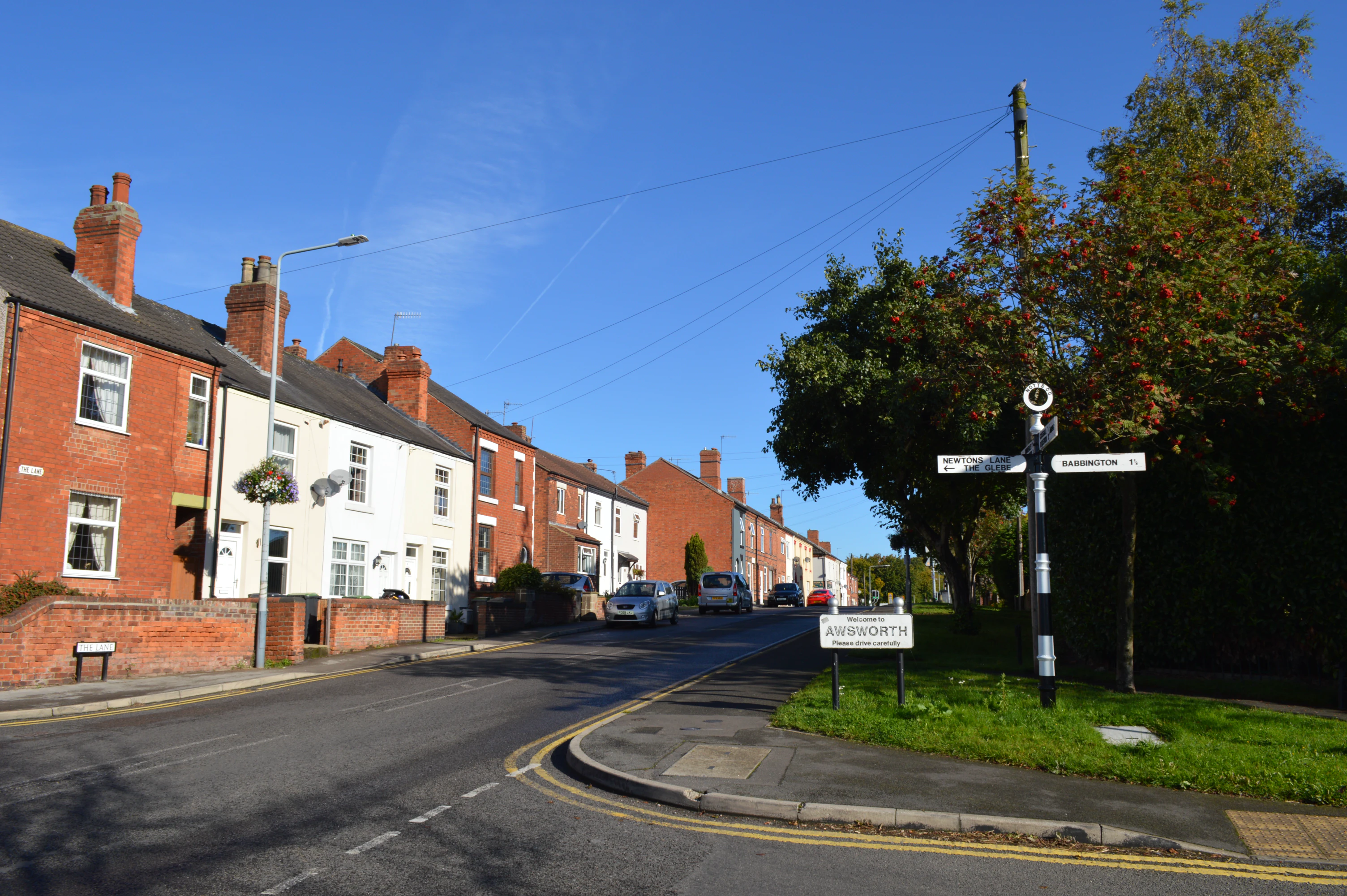 Street view in Awsworth with brick and white houses on the left and a signpost indicating Newtons Lane, The Glebe, and Babington, under a clear blue sky.