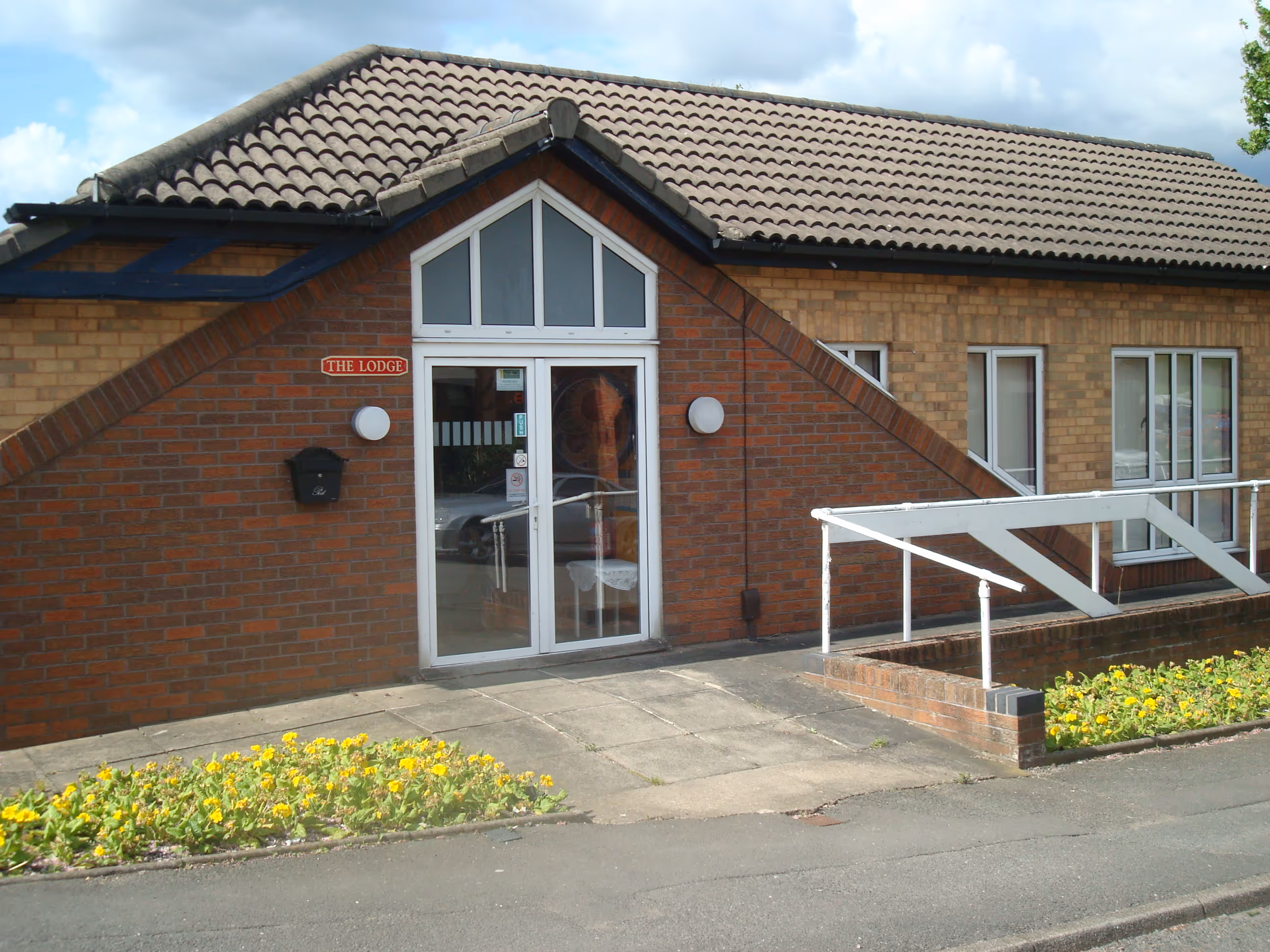 Brick building entrance with a glass door labeled 'The Lodge,' flanked by two round lights and yellow flowers along the sidewalk.