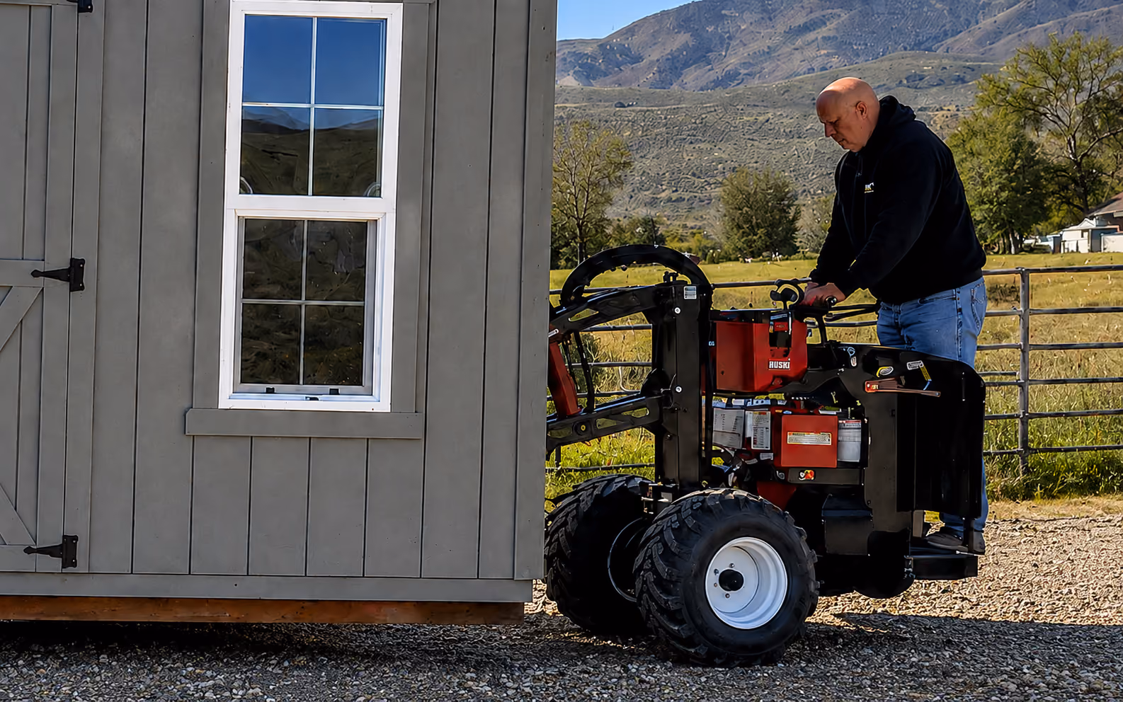 Man operating a shed-moving machine beside a gray storage shed on a gravel driveway with fields and mountains in the background.