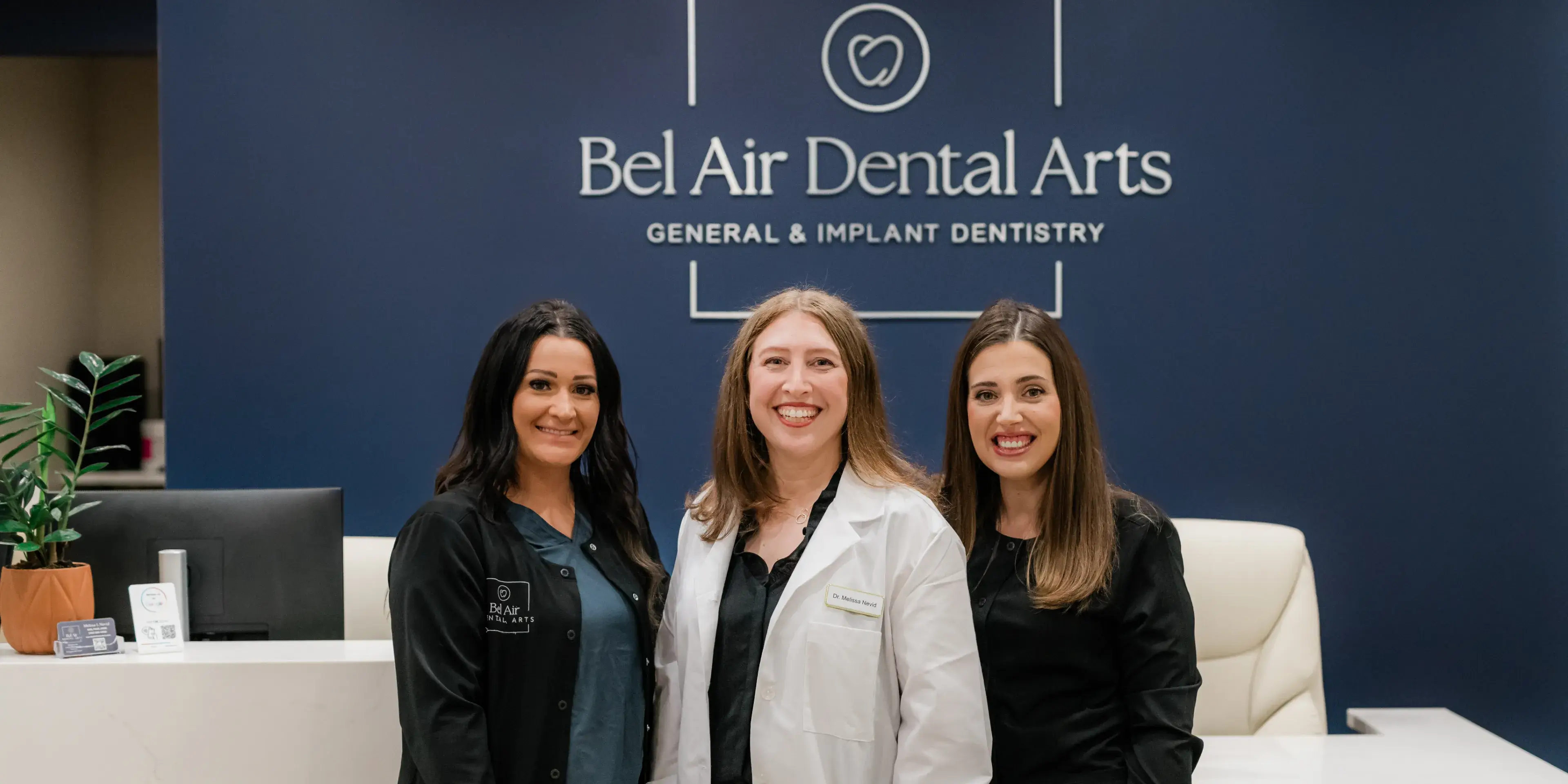 Three women smiling inside a dental office, standing in front of a reception desk.
