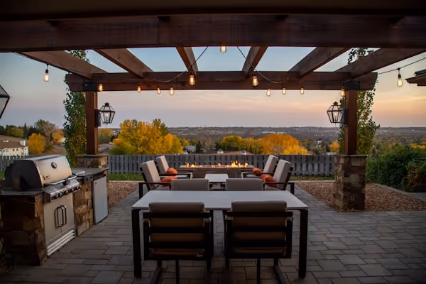 An outdoor patio area under a wooden pergola at dusk. The space has a grill, a dining table with chairs, and a seating area in the background with a fire pit. String lights hang from the pergola, and the sunset is visible over a distant landscape.