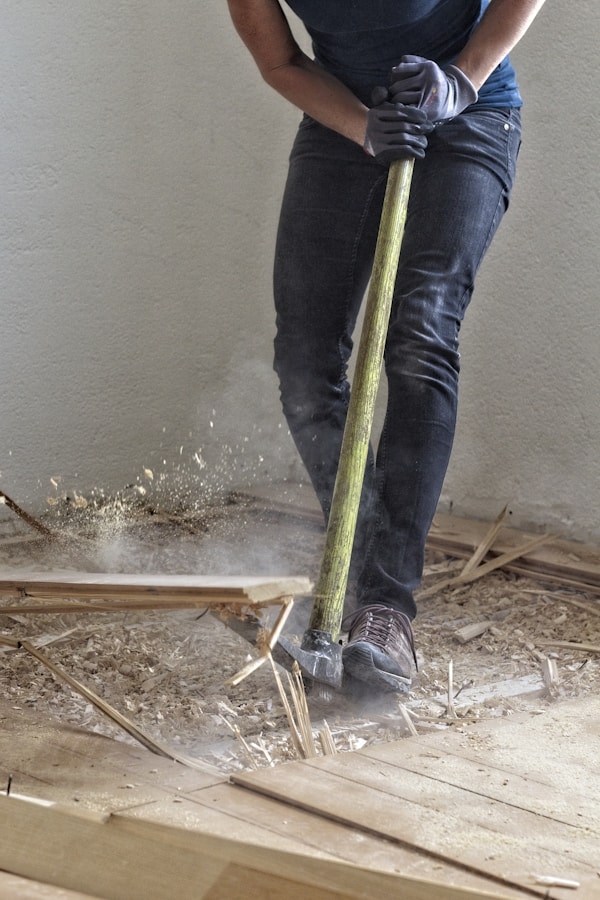 A person wearing work gloves and dark pants is using a floor scraper or pry bar to remove old wooden flooring, kicking up dust in the process. The focus is on the action, and the background is a simple wall.