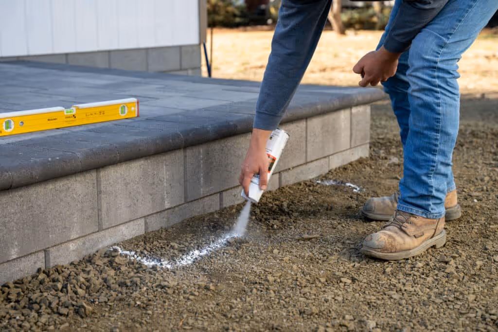 A person wearing jeans and work boots is kneeling, spraying a white powder from a can onto the gravel ground. A newly constructed patio or step made of large gray pavers is visible behind them, with a yellow level resting on it.