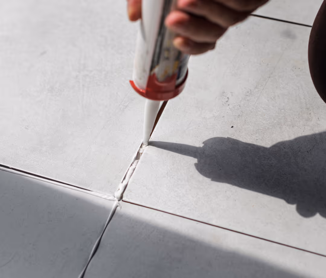 A close-up of a person's hand using a caulk gun to apply sealant to the grout lines between light-colored floor tiles. A shadow is cast from the person onto the tiles.
