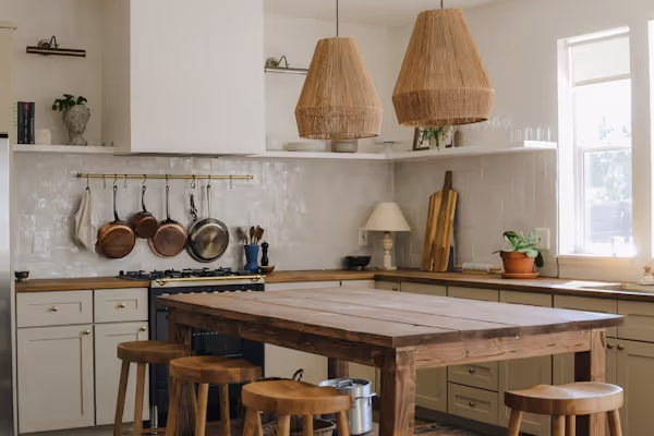 A warm, rustic kitchen with a large wooden island and four wooden stools. The kitchen features off-white cabinets, a tiled backsplash, and a hanging rack with copper pots and pans. Two large woven pendant lights hang above the island. A window lets in natural light on the right.