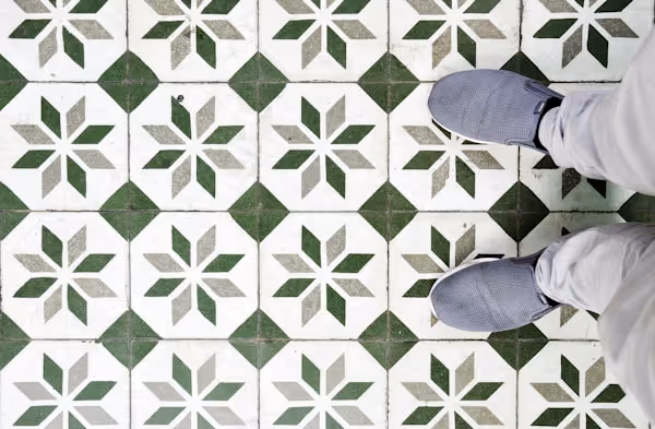 A high-angle shot shows a person's feet in light gray shoes standing on a floor with a geometric tile pattern. The tiles are white with a green and light gray starburst design, separated by solid green square tiles at the corners.