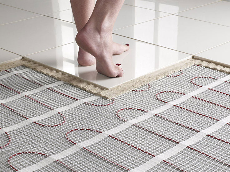 A person's bare feet are stepping onto a finished white tiled floor, which is adjacent to an unfinished area where a radiant floor heating mat with red wiring is visible. The image shows the process of installing a heated tile floor.