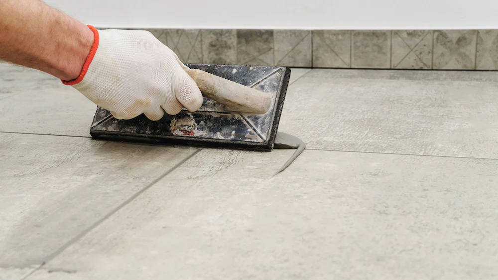A close-up of a gloved hand using a grout float to apply grout to the seams of light-colored floor tiles. The tiles have a subtle marble or stone pattern. A tiled baseboard is visible in the background.