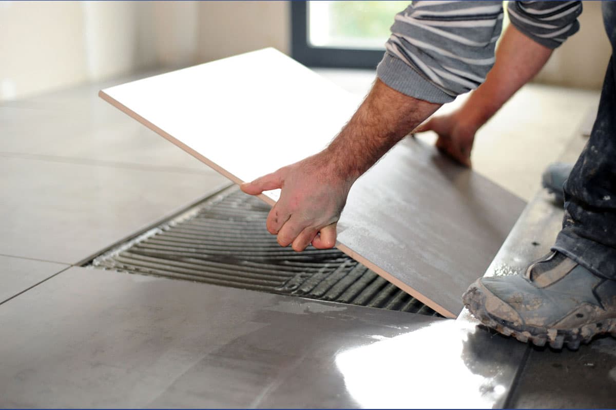 A person wearing a striped shirt and work boots is installing a large, light-colored floor tile. They are pressing the tile into a fresh bed of grooved adhesive on a floor with existing dark gray tiles. The shot is focused on the hands and the tiling process.