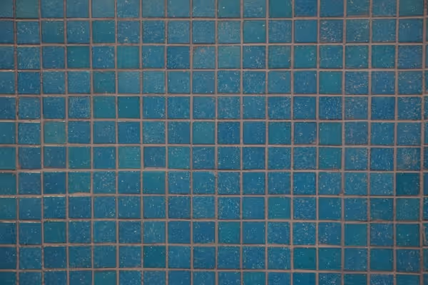 A close-up, straight-on shot of a wall or surface covered in small, square mosaic tiles. The tiles are a bright turquoise or teal color, and the grout is a darker, contrasting shade, possibly brown or gray. The texture of the tiles is slightly uneven.