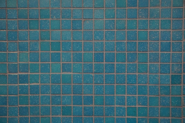 A close-up, straight-on shot of a wall or surface covered in small, square mosaic tiles. The tiles are a bright turquoise or teal color, and the grout is a darker, contrasting shade, possibly brown or gray. The texture of the tiles is slightly uneven.