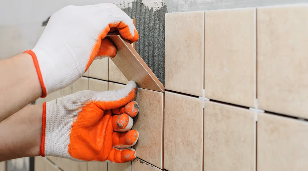 A close-up shot of gloved hands installing beige-colored wall tiles. The person, wearing white and orange work gloves, is pressing a new tile into a bed of fresh adhesive next to already placed tiles with white spacers. The wall is partially tiled.