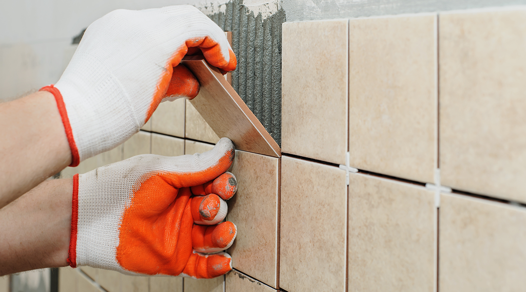 A close-up shot of gloved hands installing beige-colored wall tiles. The person, wearing white and orange work gloves, is pressing a new tile into a bed of fresh adhesive next to already placed tiles with white spacers. The wall is partially tiled.