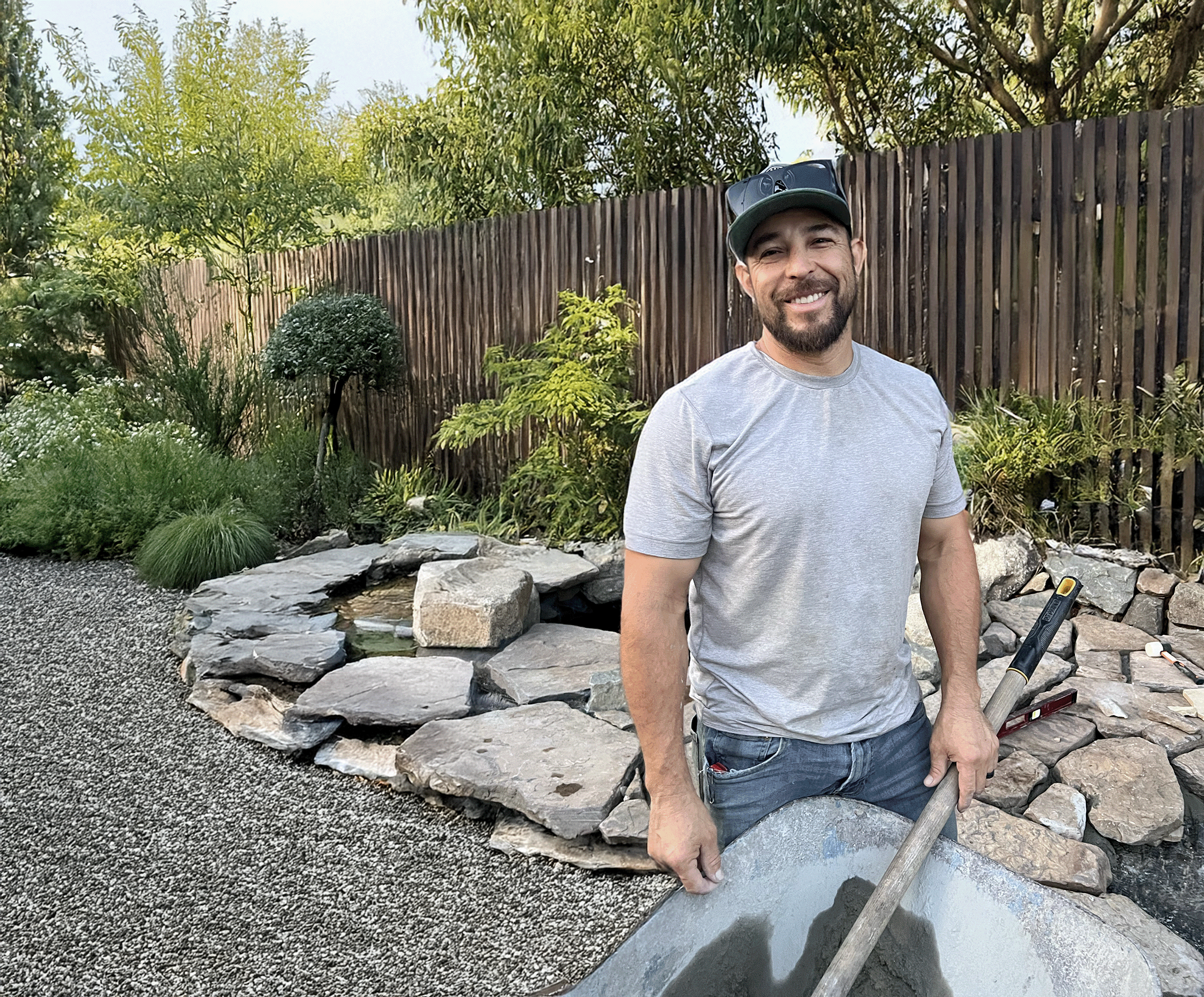 A smiling man with a beard and a baseball cap stands in a backyard. He is wearing a gray t-shirt and jeans and is holding a shovel in a wheelbarrow filled with cement or soil. Behind him is a decorative rock formation with a small pond, a wooden fence, and green foliage.