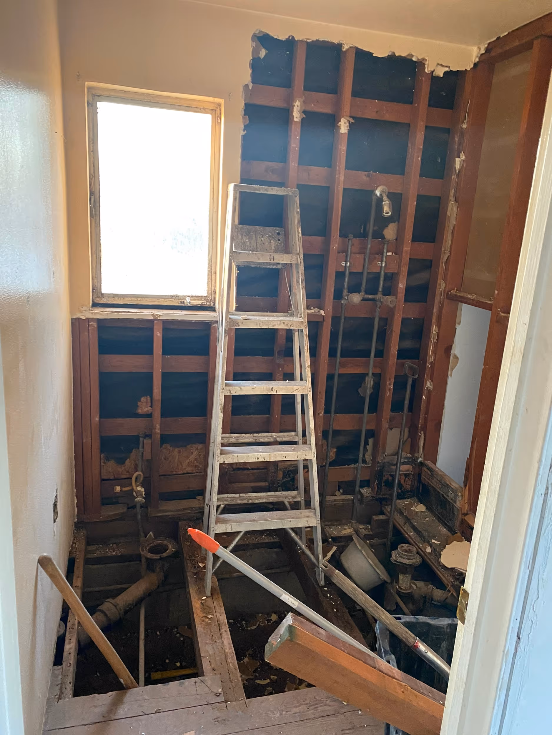 
This image documents a bathroom in the early stages of a full renovation or gut job, revealing the structural elements and plumbing. All finished surfaces have been removed, exposing the wooden wall studs and floor joists, with areas of the floor completely opened up to show the pipes and subfloor below. A metal step ladder stands prominently in the center of the debris-filled space, alongside various tools and pieces of wood. The rough condition of the room, with exposed wiring and plumbing pipes against the dark framework, clearly indicates a major construction project in progress, transforming the old space into something new.