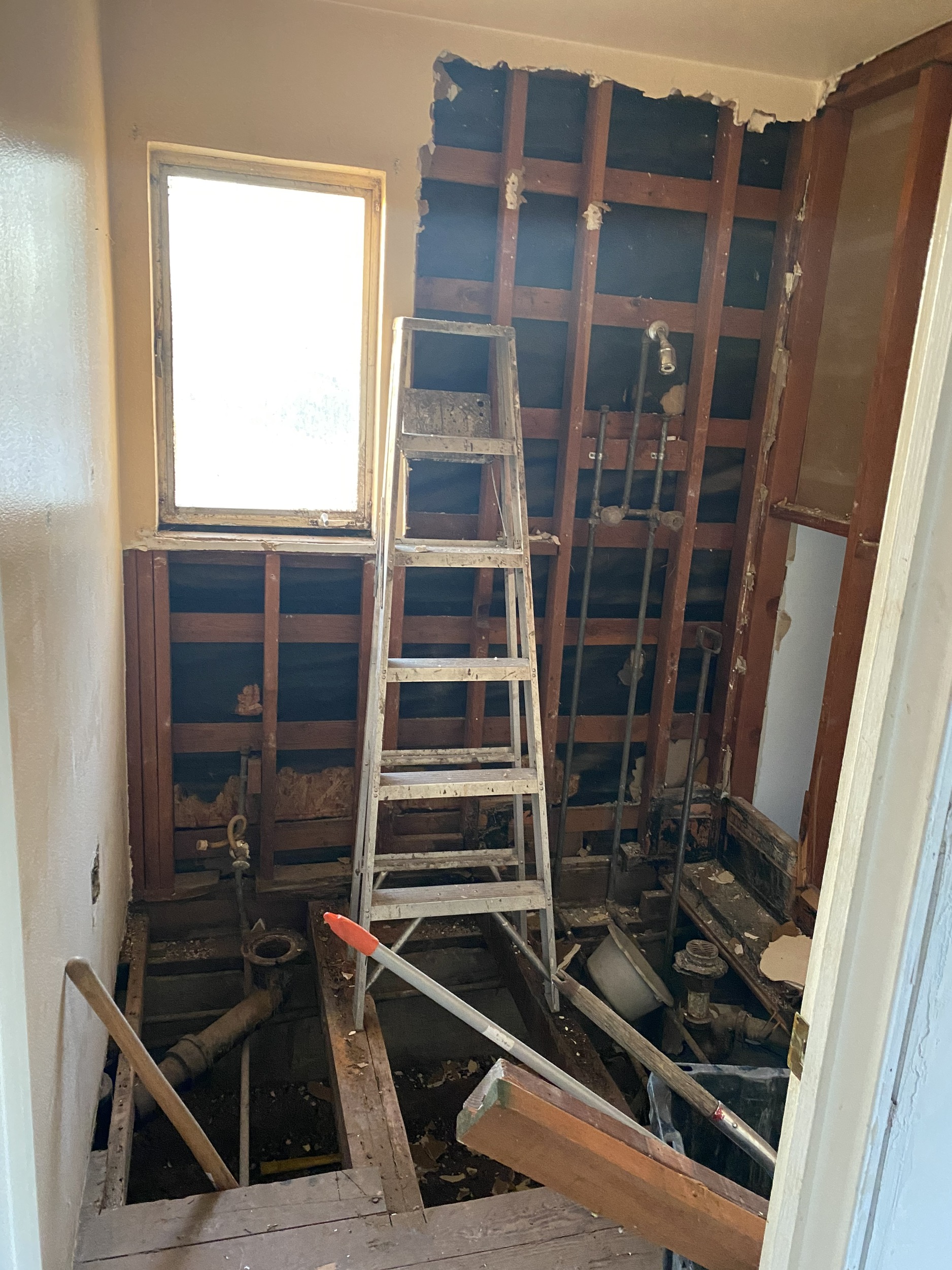 A bathroom under construction or demolition, with the floor and walls partially removed, exposing wooden studs, plumbing pipes, and a ladder standing in the center. A bright window is visible on the left. Debris and tools are scattered on the unfinished floor.