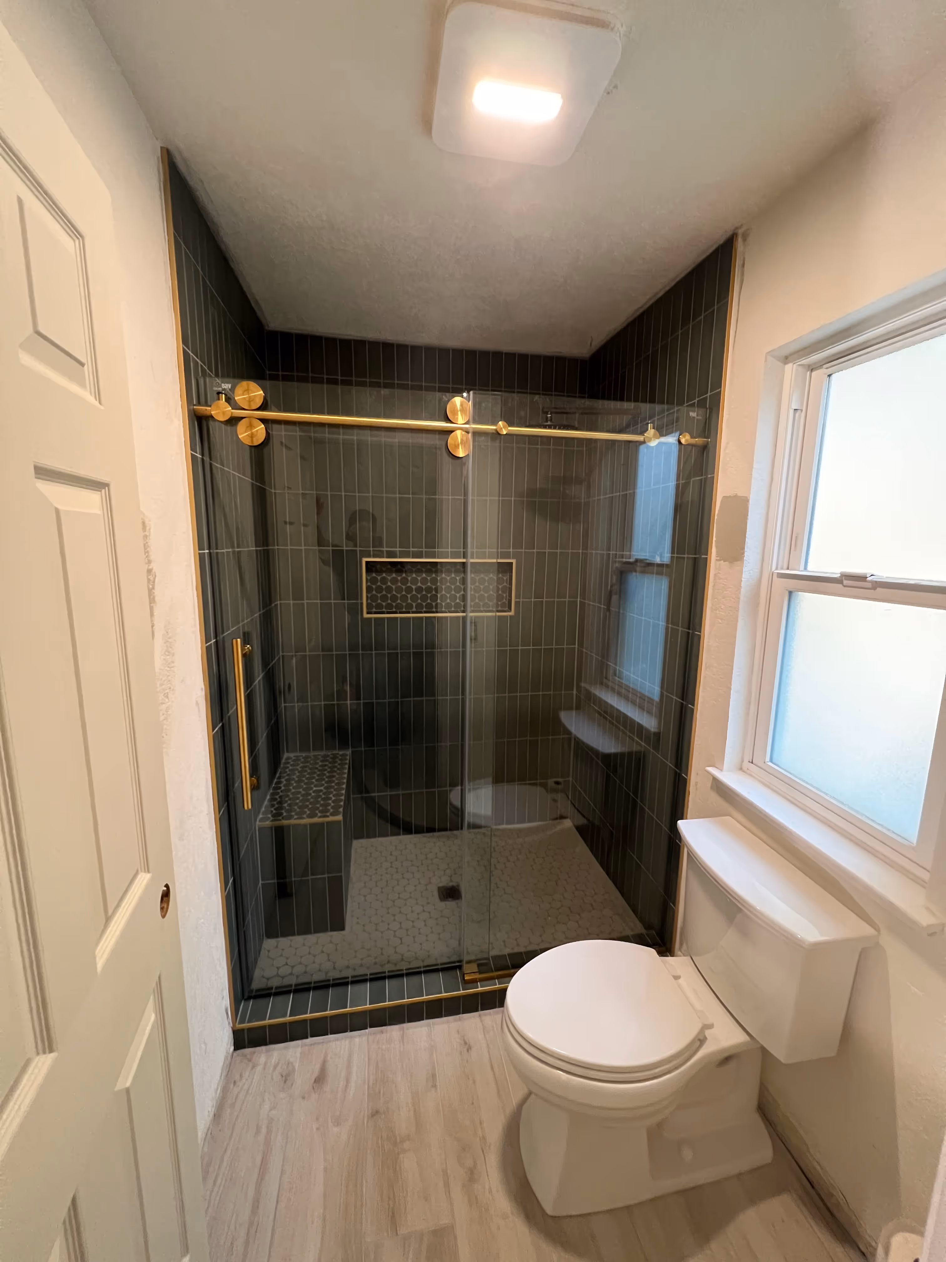 A finished, modern bathroom with a dark tiled shower enclosure and a white toilet. The shower has a glass door with a gold frame and hardware, a dark-tiled bench, and a recessed niche. The floor is light wood-effect vinyl or tile. A window is above the toilet, and a white door is on the left.
