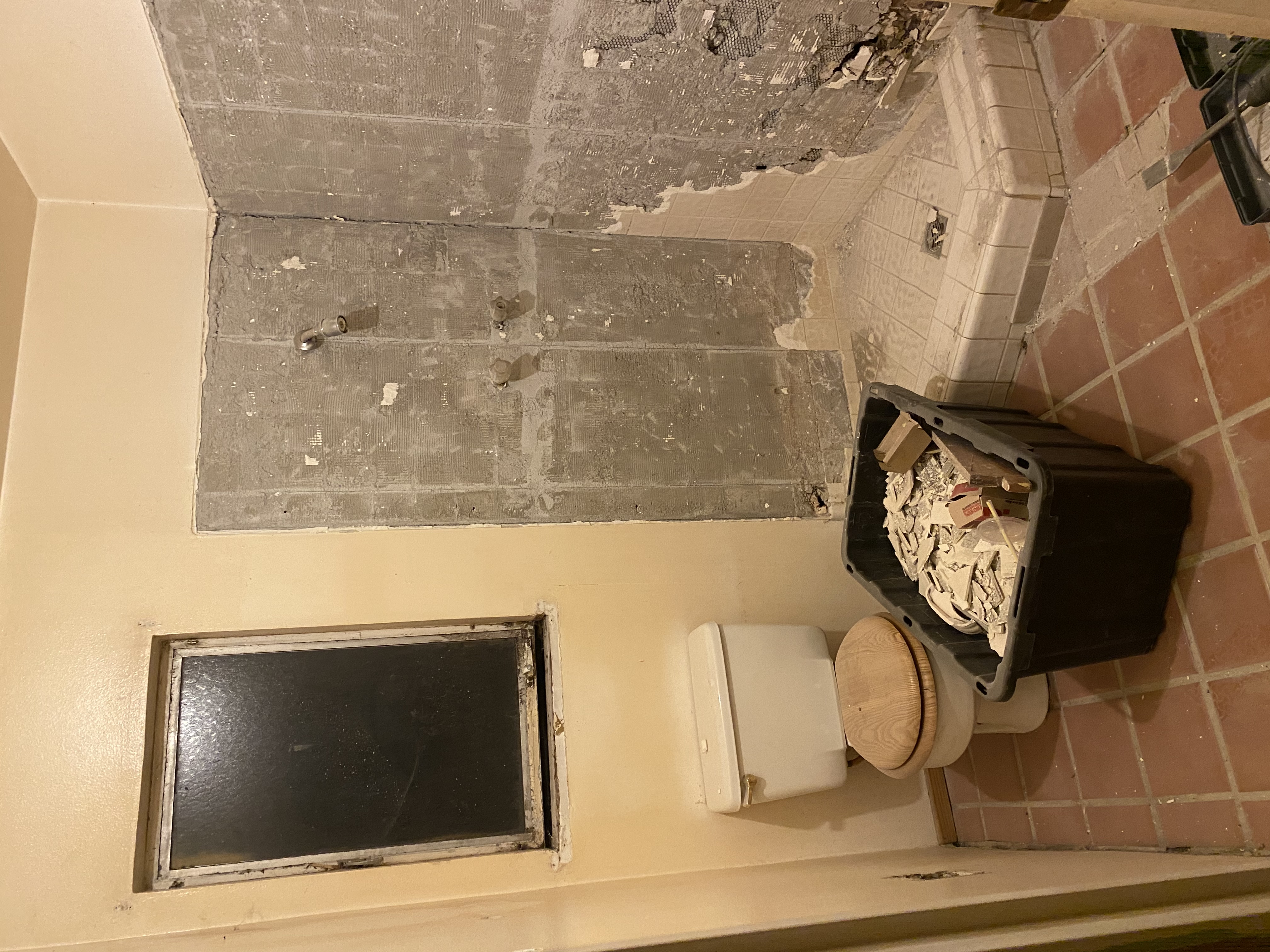 A bathroom undergoing demolition. The walls are partially torn out, exposing the underlying plaster and studs in the shower area. A toilet with a wooden seat is on the left next to a small window. A large black bin filled with debris sits on the terracotta-colored tiled floor.