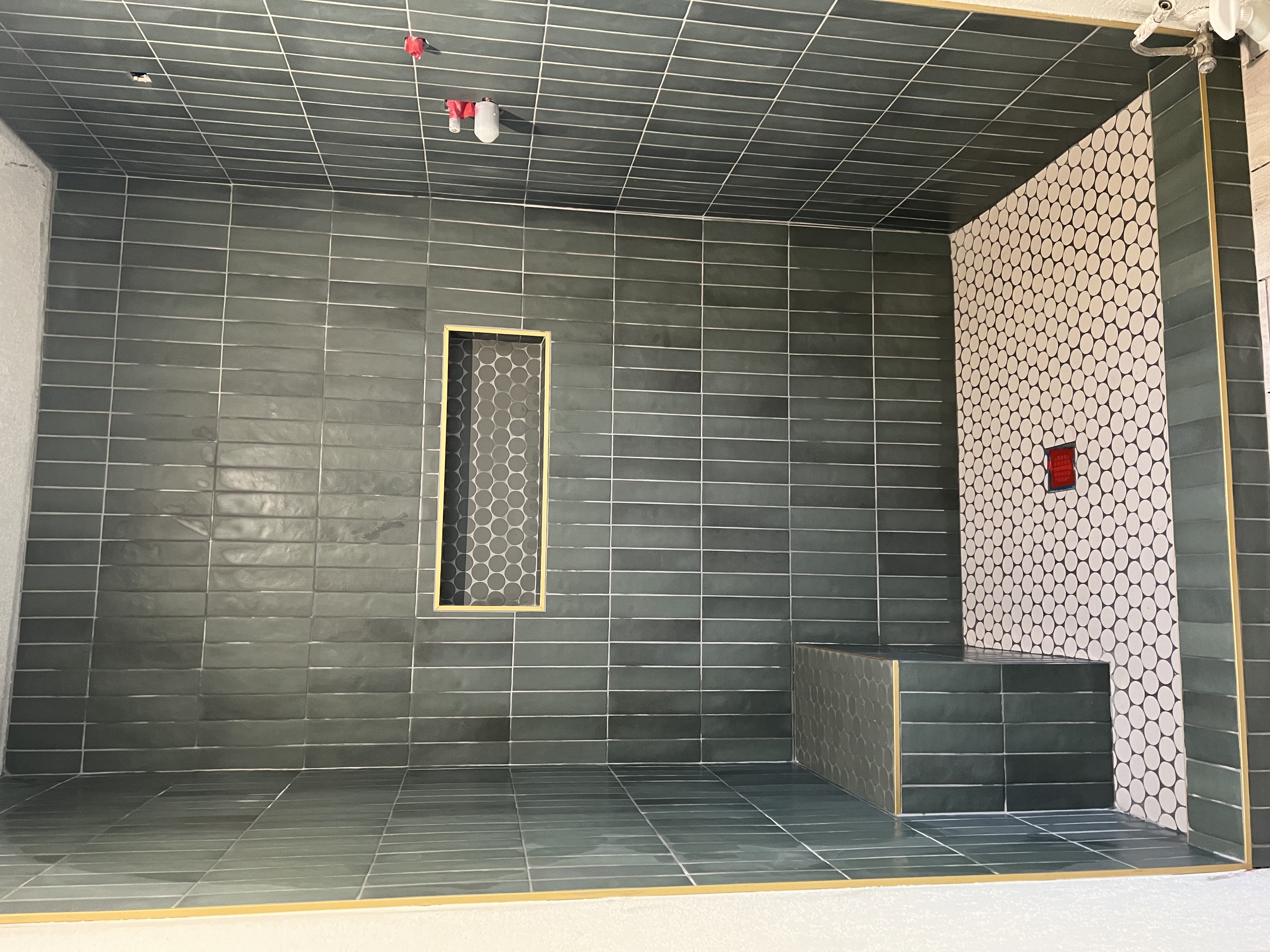 A walk-in shower with dark green vertical subway tiles on the walls and a white hexagonal tile floor. A tiled bench is on the left. The grout lines and a recessed niche have a light-colored, possibly gold, trim. A red square drain is visible in the center of the floor.