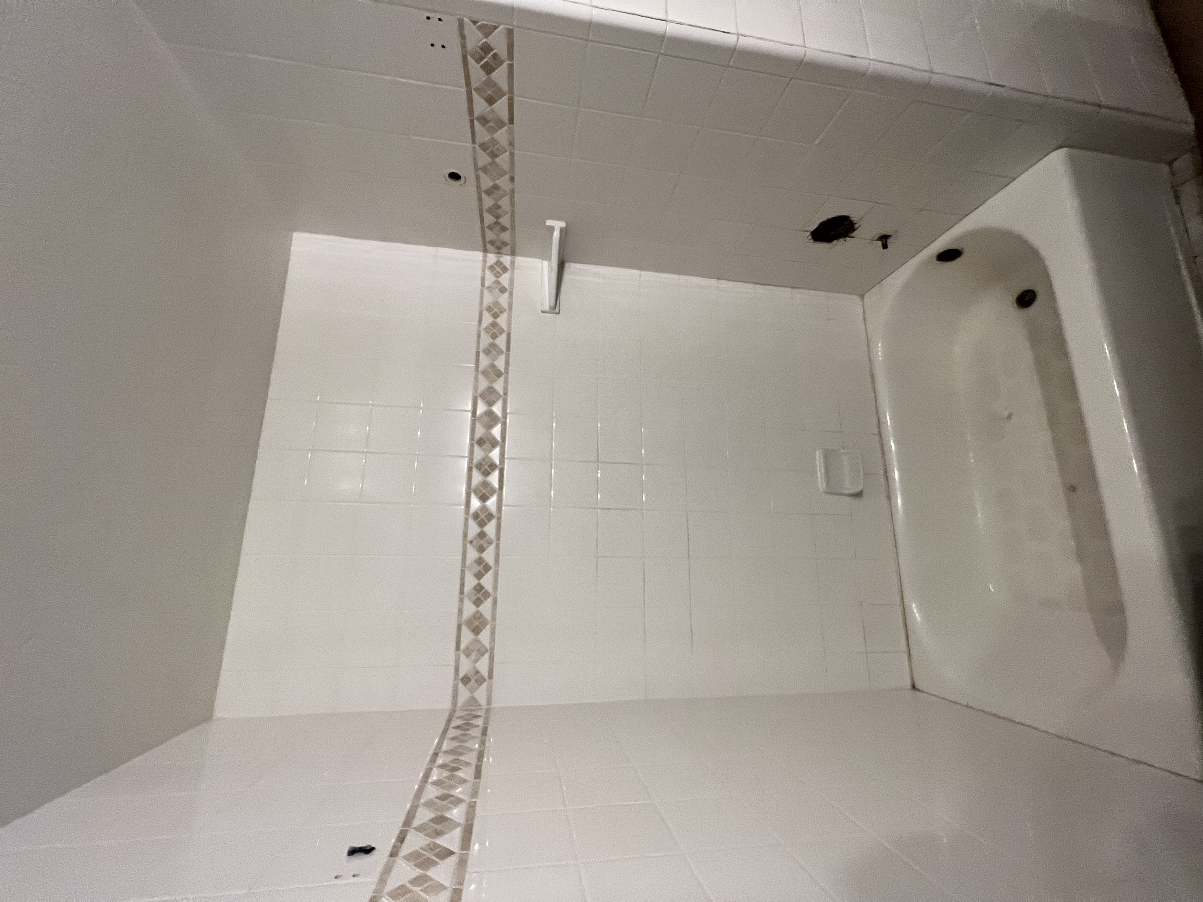 A white-tiled bathtub and shower area in need of renovation. The walls have large white square tiles, with a horizontal decorative band of smaller diamond-patterned tiles. A white soap dish and a horizontal grab bar are installed. There are small holes in the tile where fixtures have been removed.