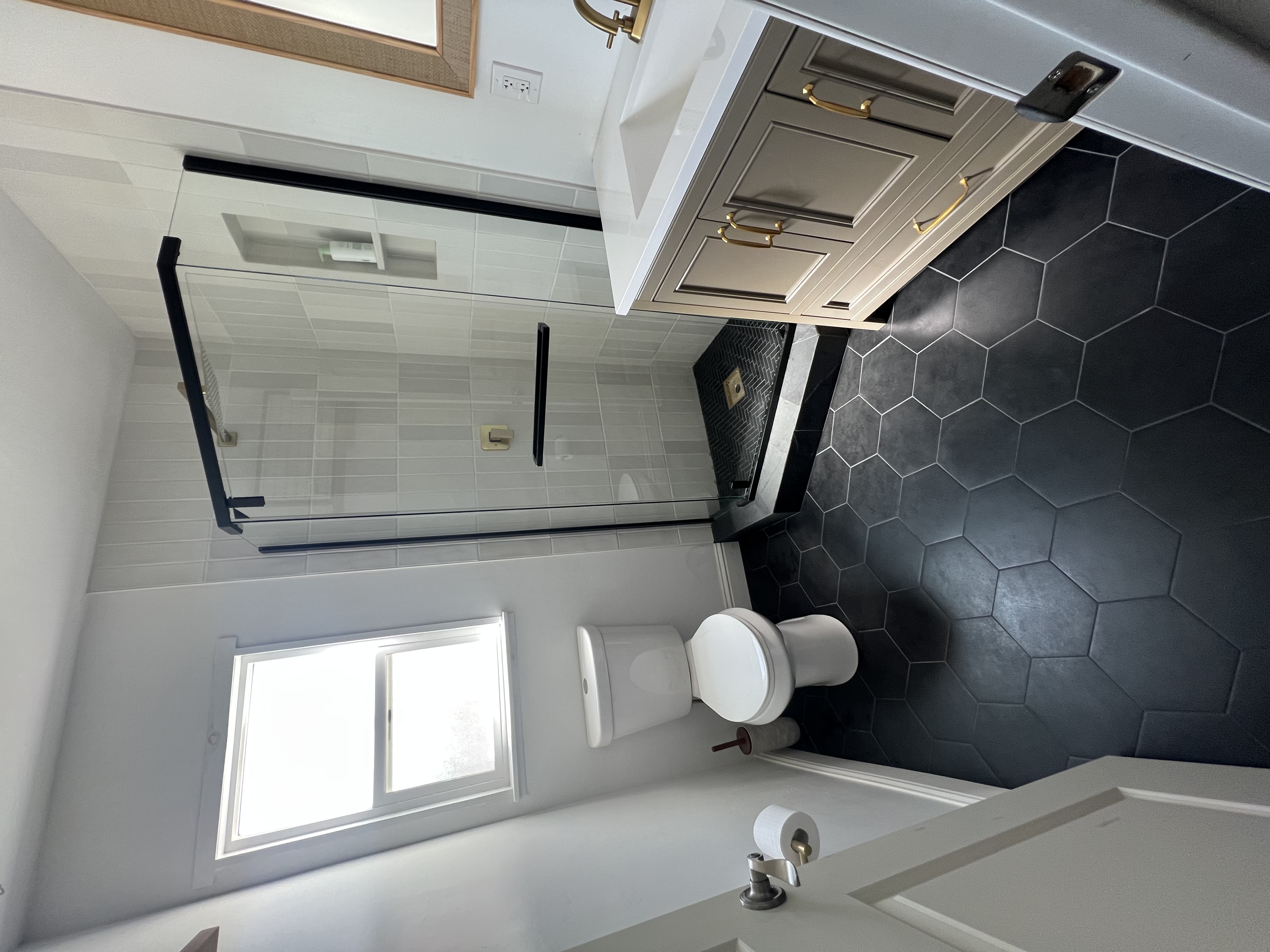 A small, modern bathroom with a dark hexagonal tile floor. The shower has a black-framed glass enclosure and vertical white tiles. A light-colored vanity with a white countertop, gold handles, and a gold faucet is on the right. A toilet is next to a window on the left.