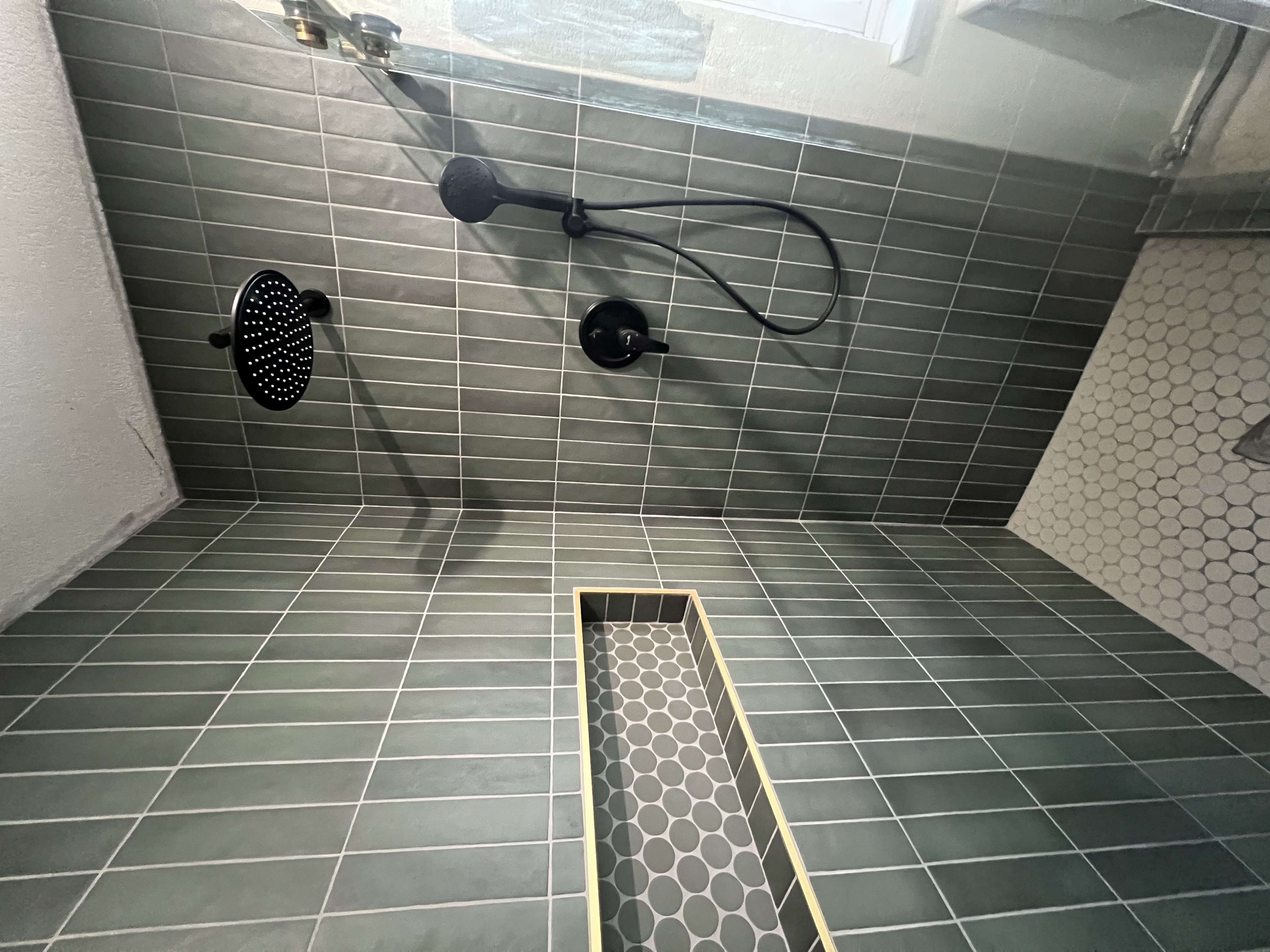 A finished shower interior with dark vertical subway tiles on the walls and a white hexagonal tile floor. A black showerhead and handheld sprayer are installed. A recessed niche with a matching hexagonal tile pattern is visible, trimmed in gold or light wood.