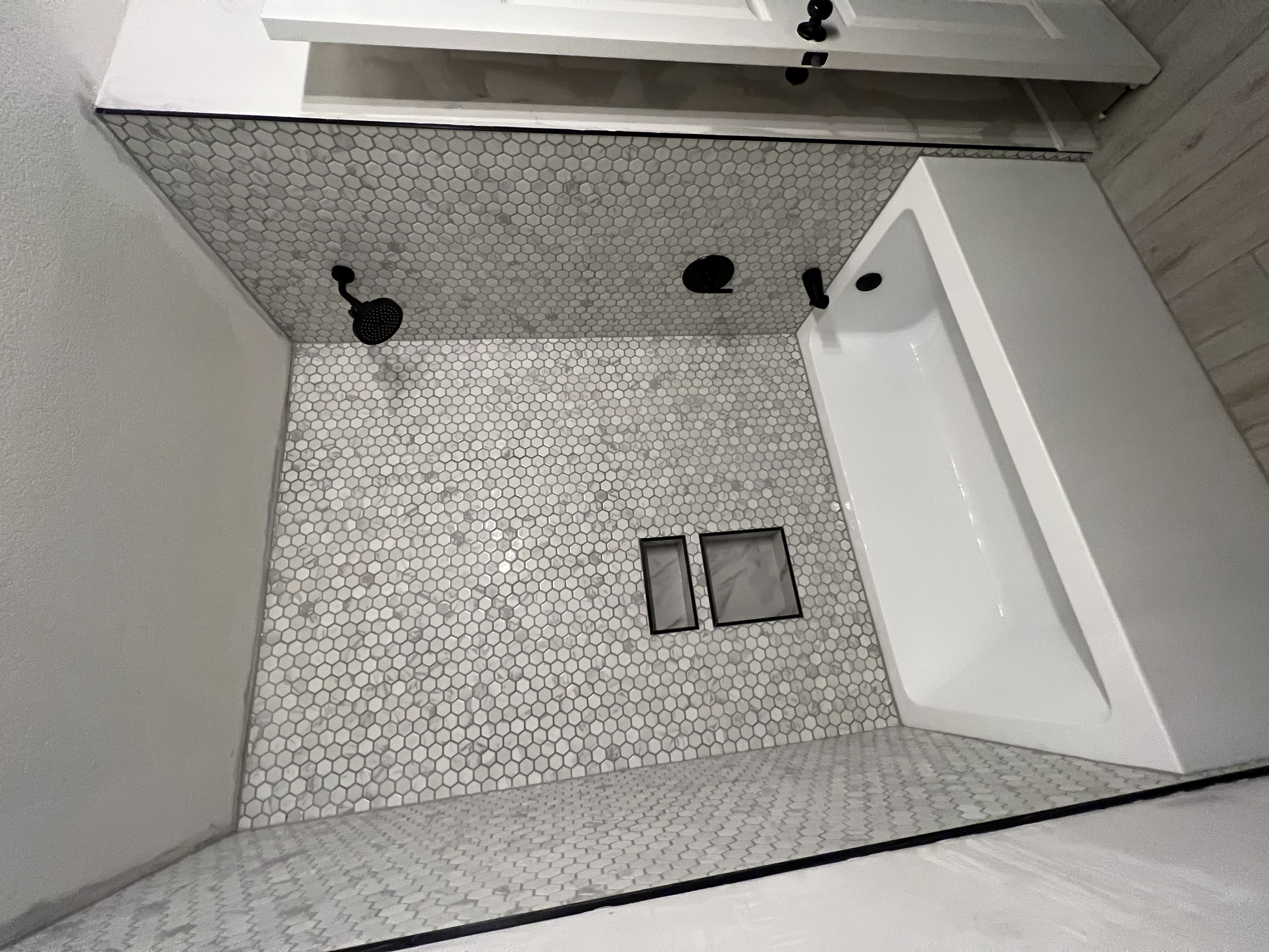 A renovated bathroom with a white bathtub and walls covered in light-colored hexagonal tiles. The shower fixtures, including a showerhead, faucet, and bath spout, are all matte black. Two recessed niches with darker tile are visible in the wall. The floor is a light wood-effect tile.