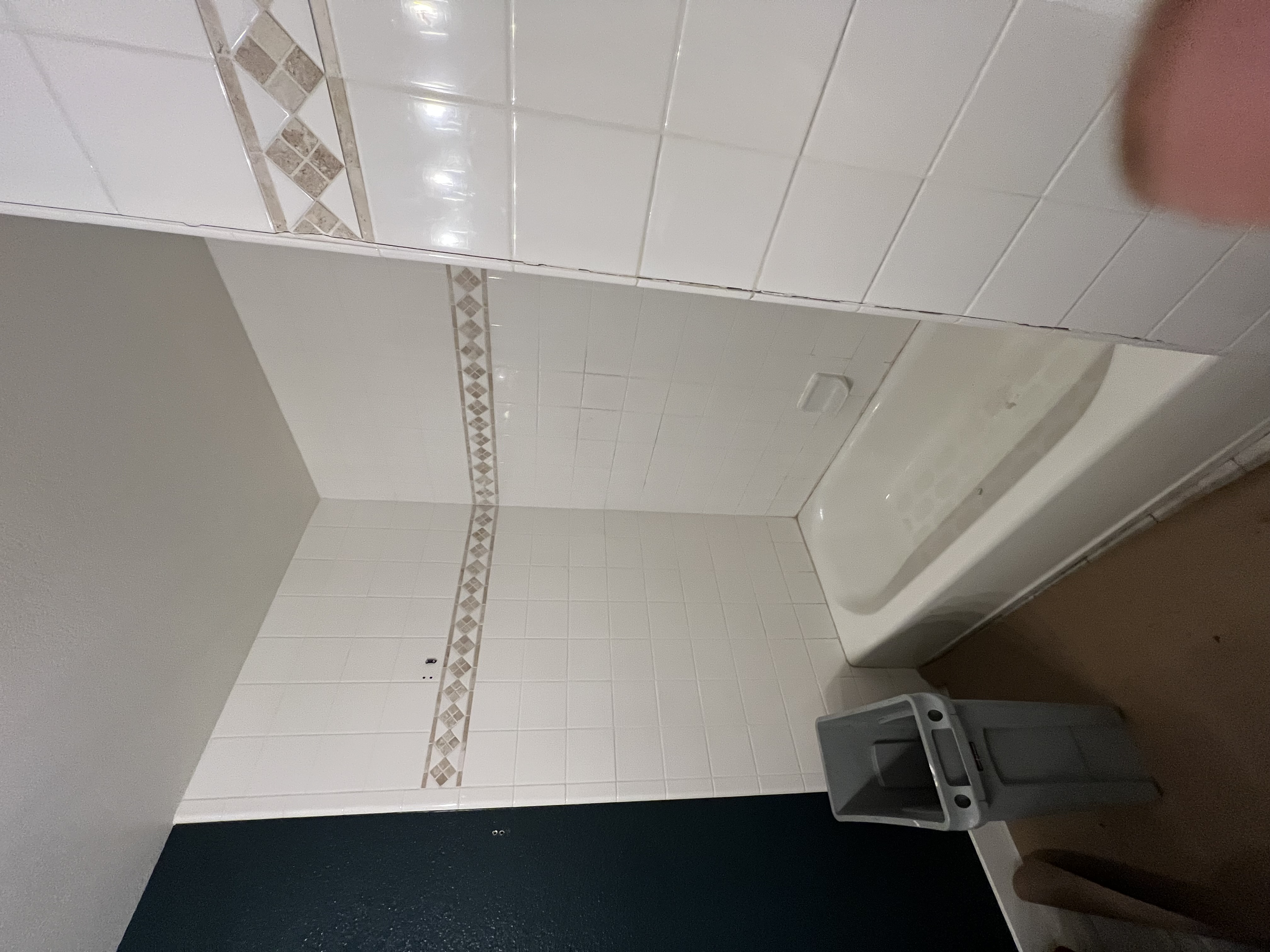 A small bathroom with an old white bathtub and white square tiles on the shower walls. A decorative tile band with a diamond pattern runs horizontally across the walls. A small gray bin sits on a carpeted floor. A dark green wall is visible on the left.