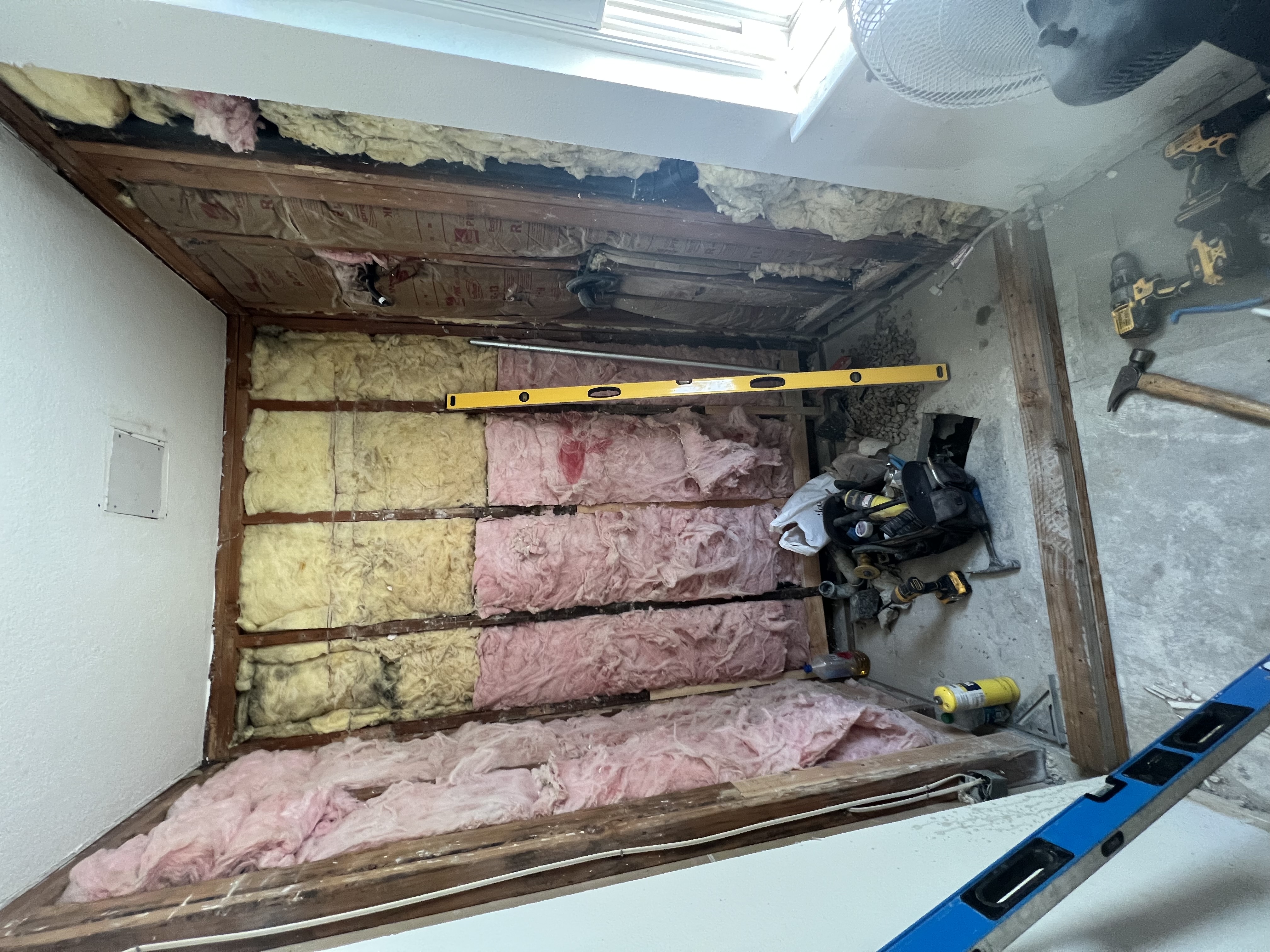 A room under renovation, with walls and floor removed to expose wooden studs and insulation. A yellow-and-black level, tools, and a drill are on the floor. Pink and yellow insulation is visible in the walls, with a small shower pan visible on the floor.