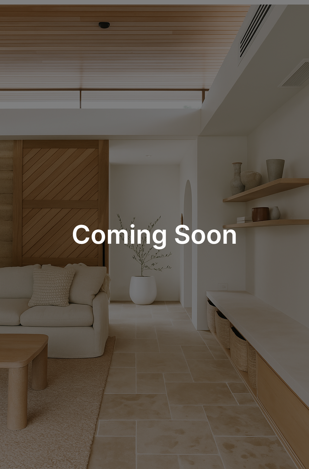 The image shows a clean, modern interior with a white couch, a wooden coffee table, and light-colored tiled floor. A wooden slatted door and floating shelves are visible. The text "Coming Soon" is overlaid in large, white letters in the center of the image.