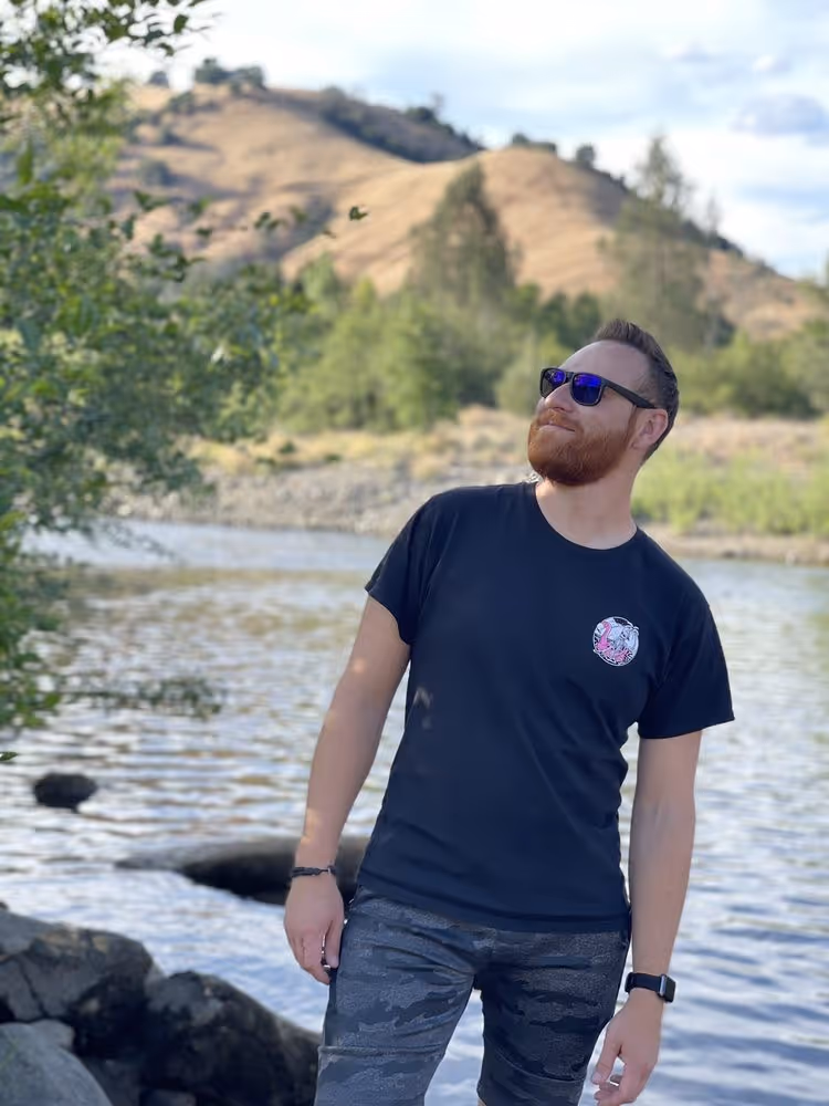A man with a reddish beard and sunglasses stands by a body of water, looking up and to his right. He is wearing a black t-shirt with a small graphic on the chest, camouflage shorts, a watch, and bracelets. In the background, there are rolling hills, green trees, and a river. The weather appears sunny with some clouds in the sky.